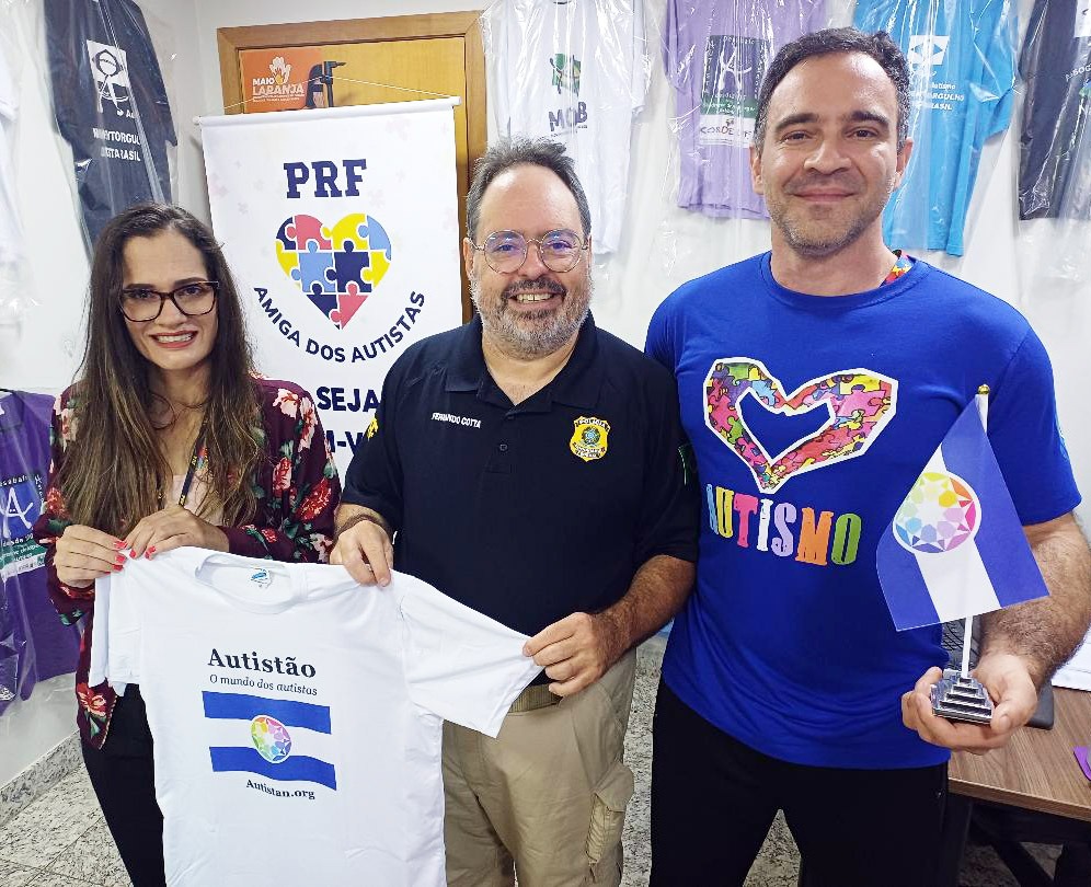 Dra. Larissa Argenta, Inspetor Fernando Cotta e Paulo Borges no escritório da PRF Amiga dos Autistas, segurando uma camiseta e a bandeira do Autistão.
